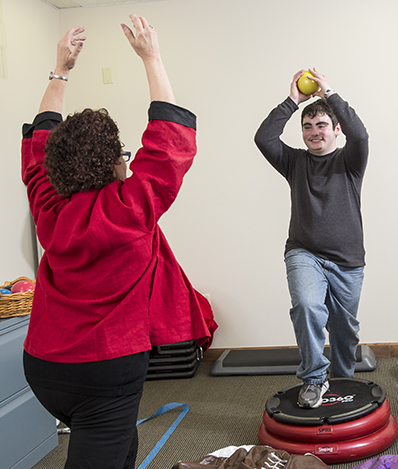 Adult is holding a small yellow ball over his head, smiling at the therapist making a similar stance